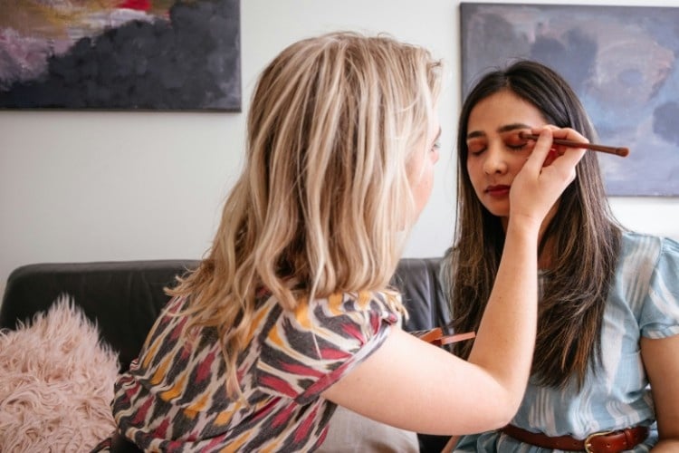 a makeup artist applies a young woman's eyeshadow. the two women are sitting on a couch